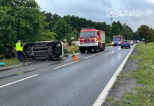 Na drodze leży przewrócony samochód, obok pracują strażacy i policjant, a w tle widać inne pojazdy.