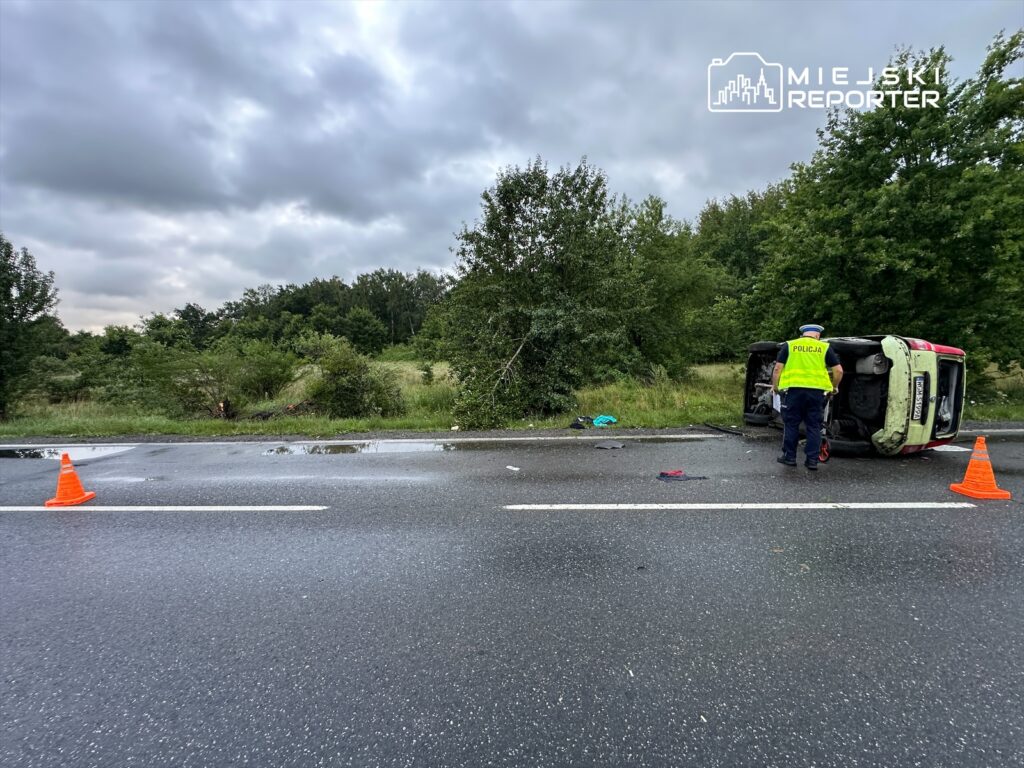 Policjant w kamizelce odblaskowej bada przewrócone na bok auto na drodze, otoczone przez pomarańczowe trójkąty ostrzegawcze.