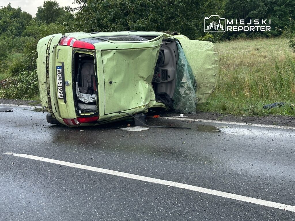 Zielony samochód osobowy leży na dachu na poboczu drogi, obok widoczna zniszczona karoseria i rozbite szkło.
