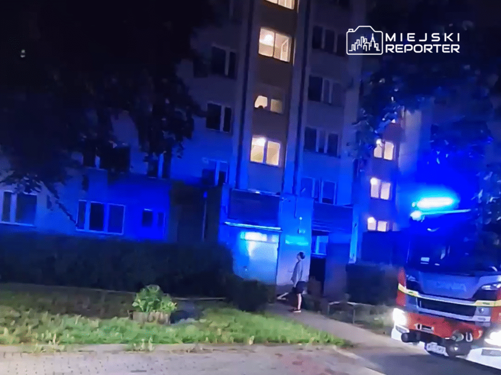 Mężczyzna w szarej bluzie stoi obok wejścia do budynku, a w tle widać wozem strażackim z niebieskimi światłami.