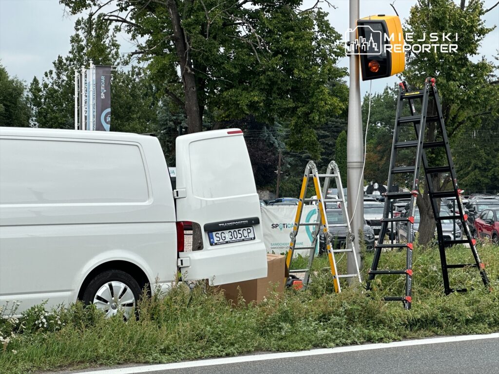 Biały furgon z otwartymi drzwiami stoi obok sygnalizacji świetlnej, a obok znajdują się drabiny i karton.