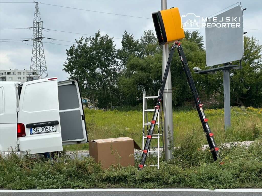 Mężczyzna przy białym vanie ustawia pomarańczowy sygnalizator na wysokim słupie obok drogi.