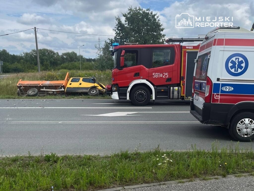 Czerwony wóz strażacki oraz ambulans zatrzymane obok uszkodzonego żółtego pojazdu na poboczu drogi.