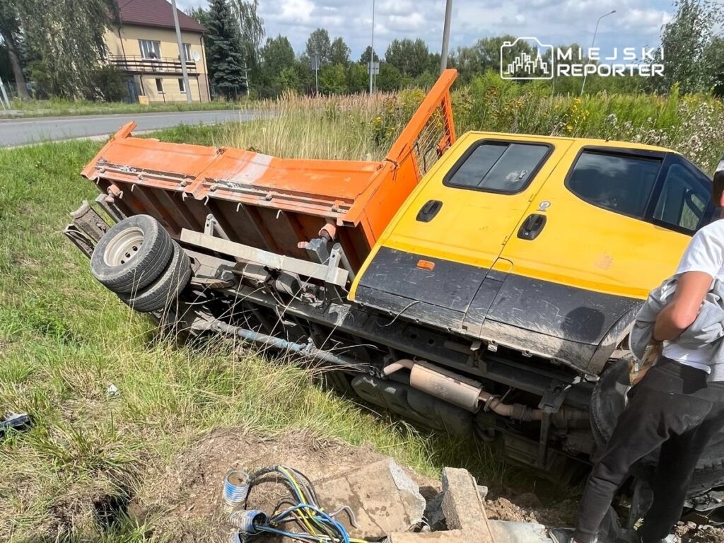 Żółty pojazd ciężarowy z pomarańczowym osprzętem leży na boku w trawie przy drodze, obok stoi mężczyzna w koszulce.