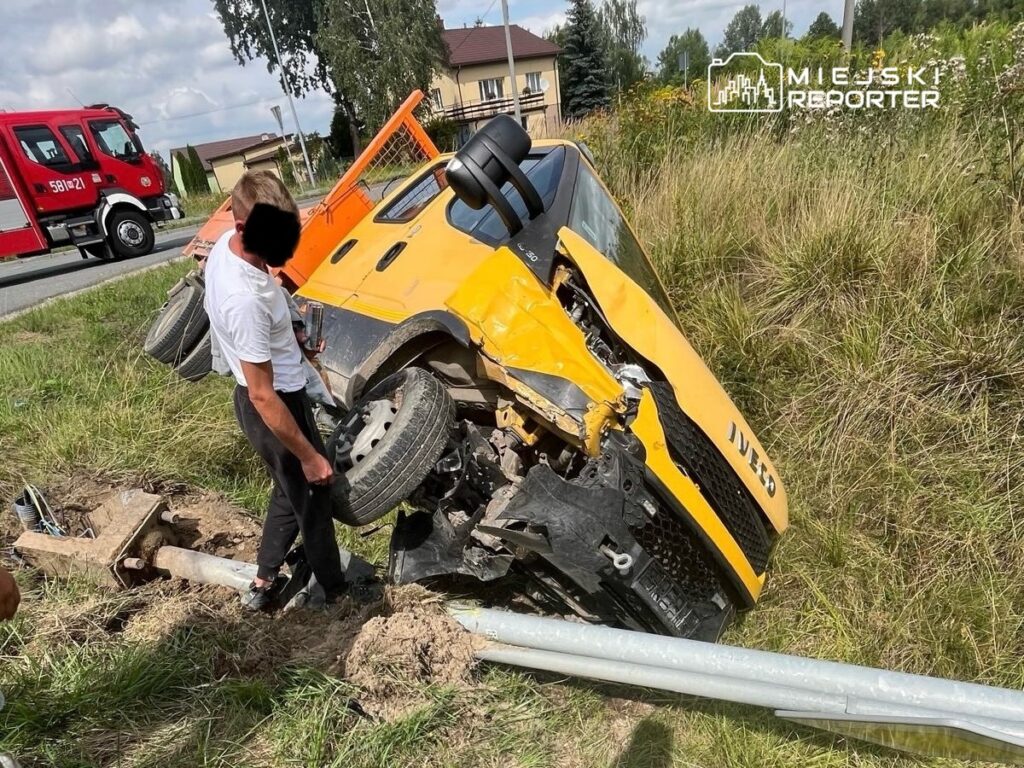 Mężczyzna w białej koszuli sprawdza przewrócony żółty pojazd na poboczu drogi, obok widoczny wóz strażacki.