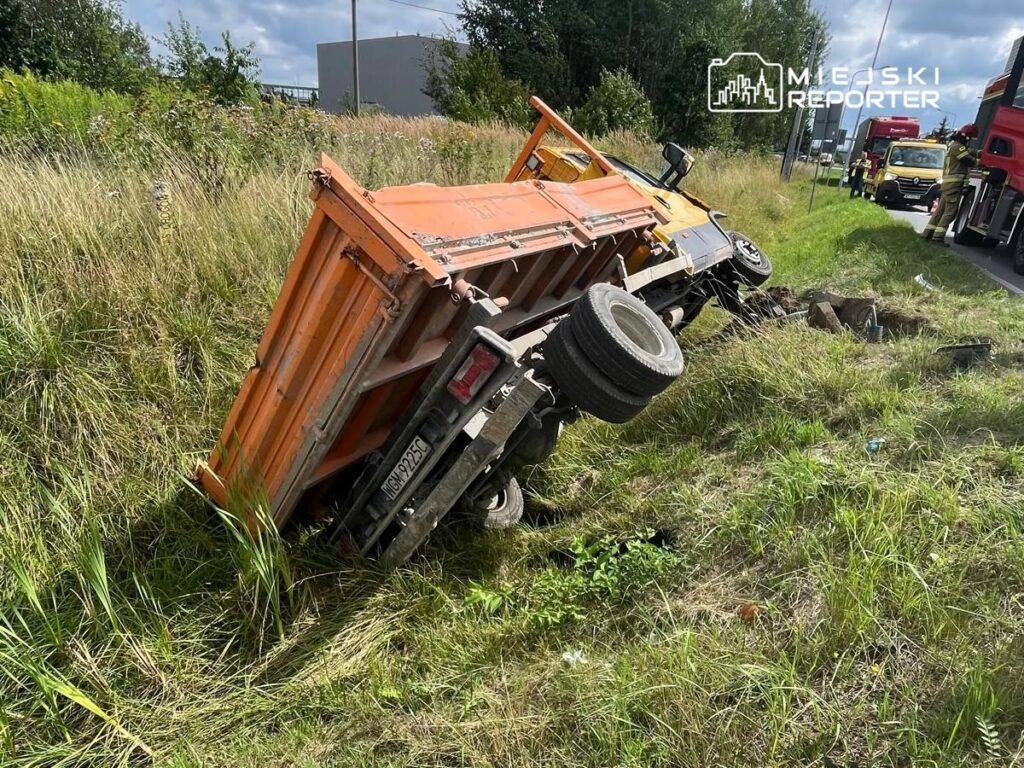 Złomowany pomarańczowy samochód ciężarowy leży na boku w wysokiej trawie przy drodze, obok stoją inne pojazdy.