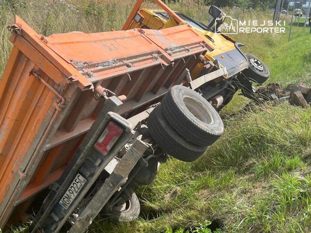 Pomarańczowa ciężarówka leży na boku w rowie przy drodze, z widocznymi uszkodzeniami i porośniętą trawą okolicą.