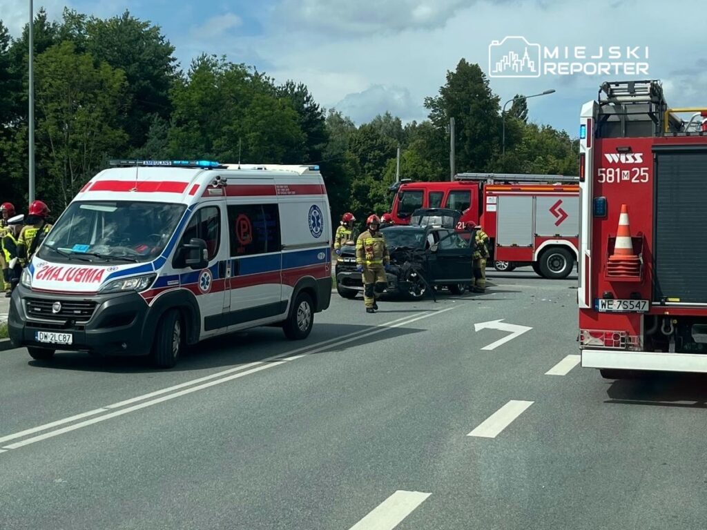 Karetka pogotowia oraz strażacy pracują przy rozbitym samochodzie na drodze, otoczeni przez zieleń.