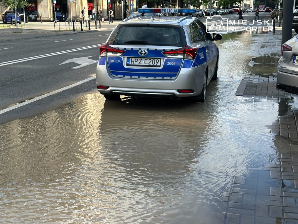 Policyjny samochód zaparkowany na ulicy obok zalanej chodnika wodą, w tle widoczne sklepy i przechodnie.