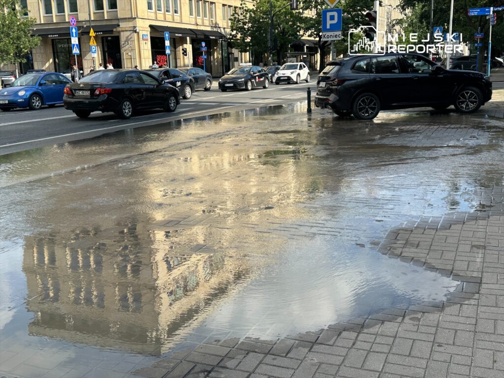 Woda zbierająca się na ulicy odbija budynki i ruch samochodów w tle, tworząc efekt lustrzanego odbicia.