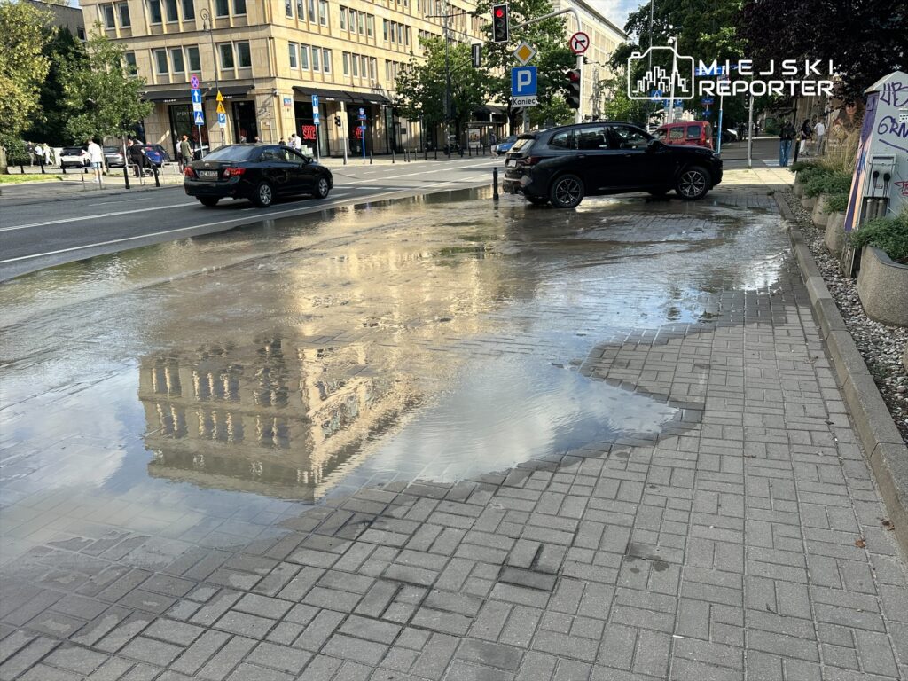 Czarna limuzyna parkująca obok kałuży wody, odbijającej budynek w tle, na ulicy w miejskim otoczeniu.