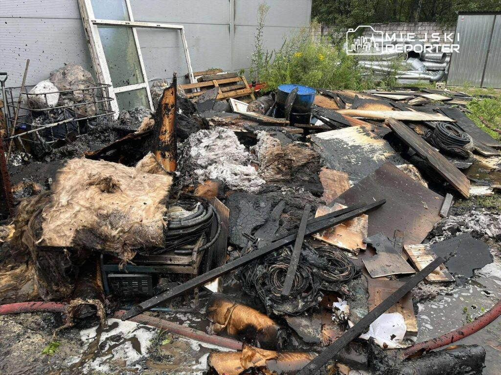 Zgliszcza po pożarze, zniszczone materiały budowlane i resztki sprzętu w nieuporządkowanej przestrzeni.