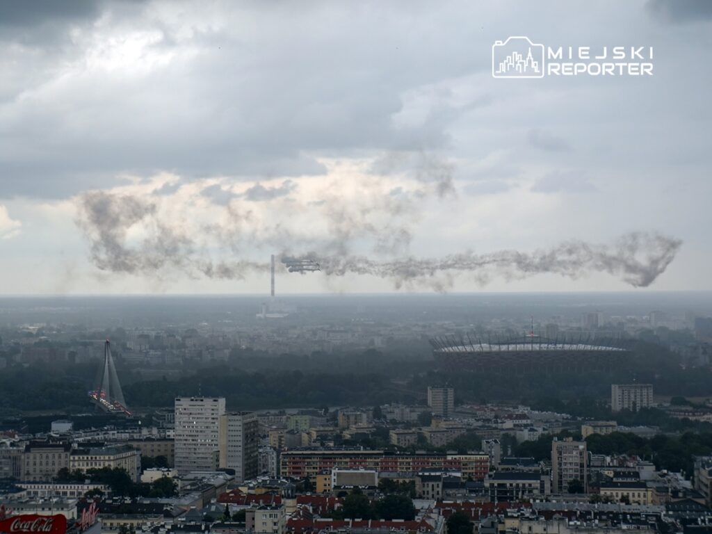 Samolot wykonujący akrobacje nad miastem, za nim widoczny stadion i chmury dymu w tle.