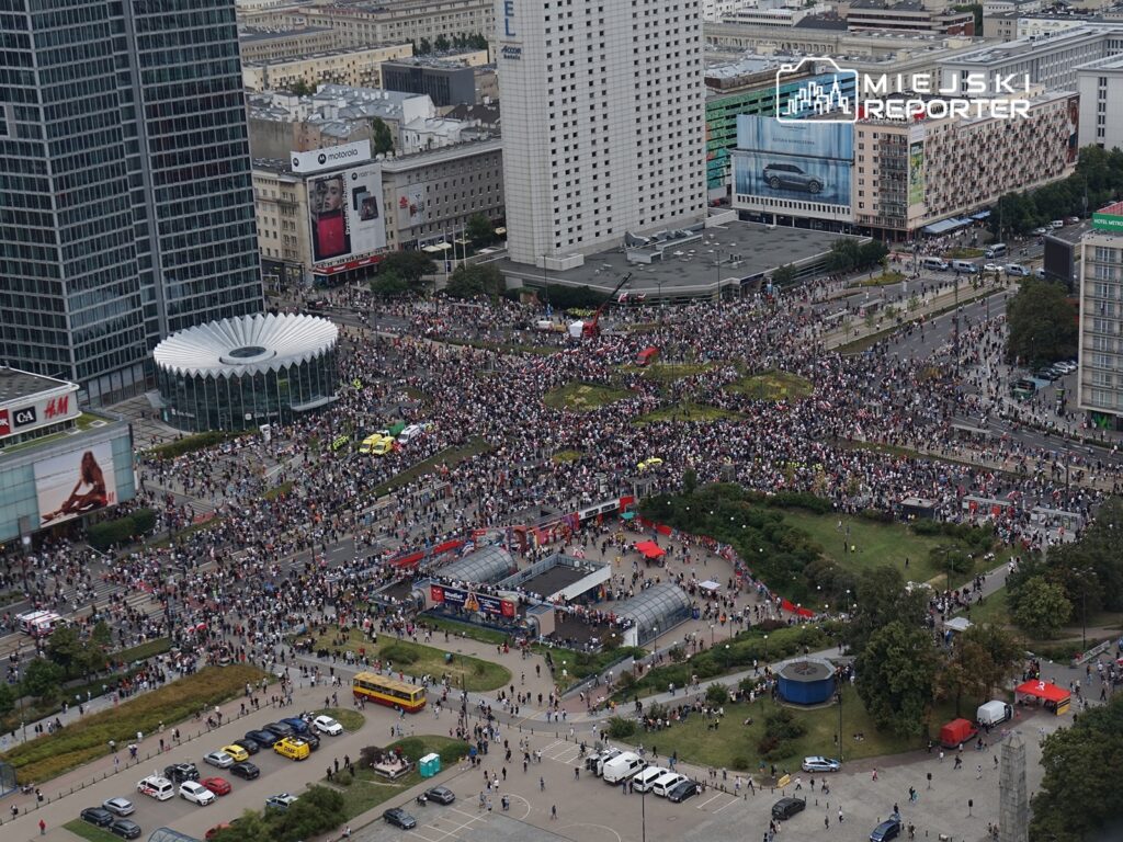 Tłum ludzi uczestniczy w demonstracji na dużym placu otoczonym nowoczesnymi budynkami.