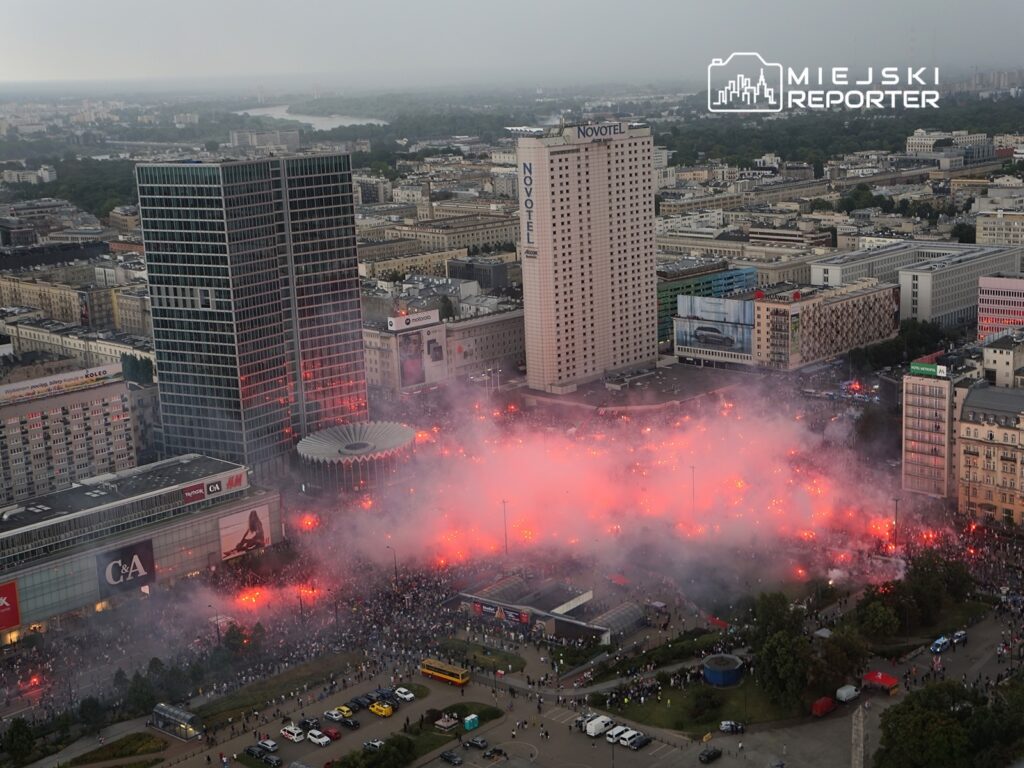 Tłum ludzi zgromadzony na placu, wypełnionym dymem i czerwonymi światłami, otoczony nowoczesnymi budynkami.