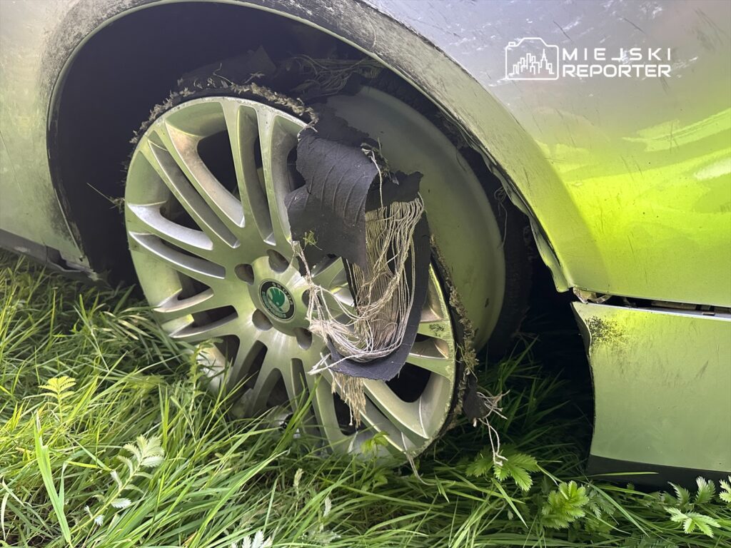 Uszkodzone koło samochodu osobowego z zerwaną oponą, otoczone wysoką trawą.