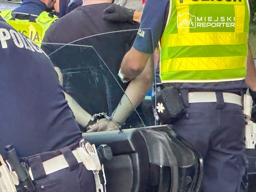 Mężczyzna z rękami w kajdankach, otoczony przez policjantów, stoi obok radiowozu w miejskim otoczeniu.