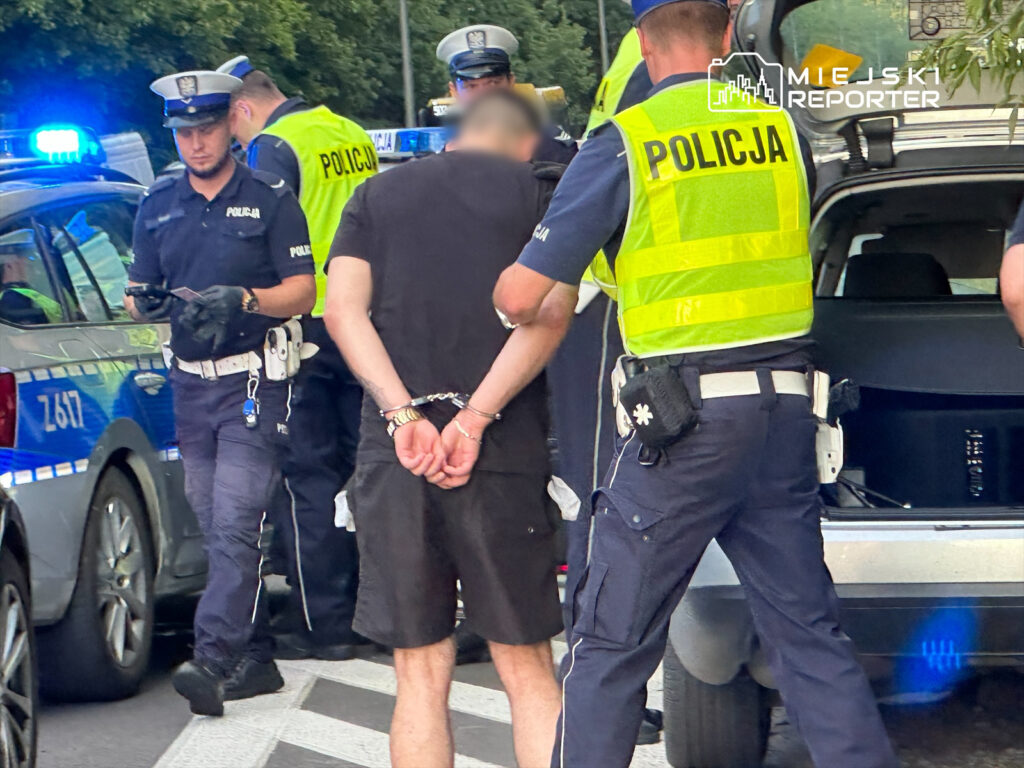 Policjanci w mundurach zatrzymują mężczyznę w kajdankach na ulicy, w tle radiowóz z włączonymi sygnałami świetlnymi.