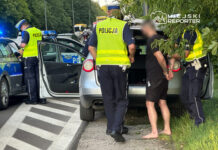 Mężczyzna w czarnej koszulce i szortach z rękami skrępowanymi przez policję stoi obok otwartego samochodu. Policjanci w kamizelkach odblaskowych sprawdzają dokumenty.
