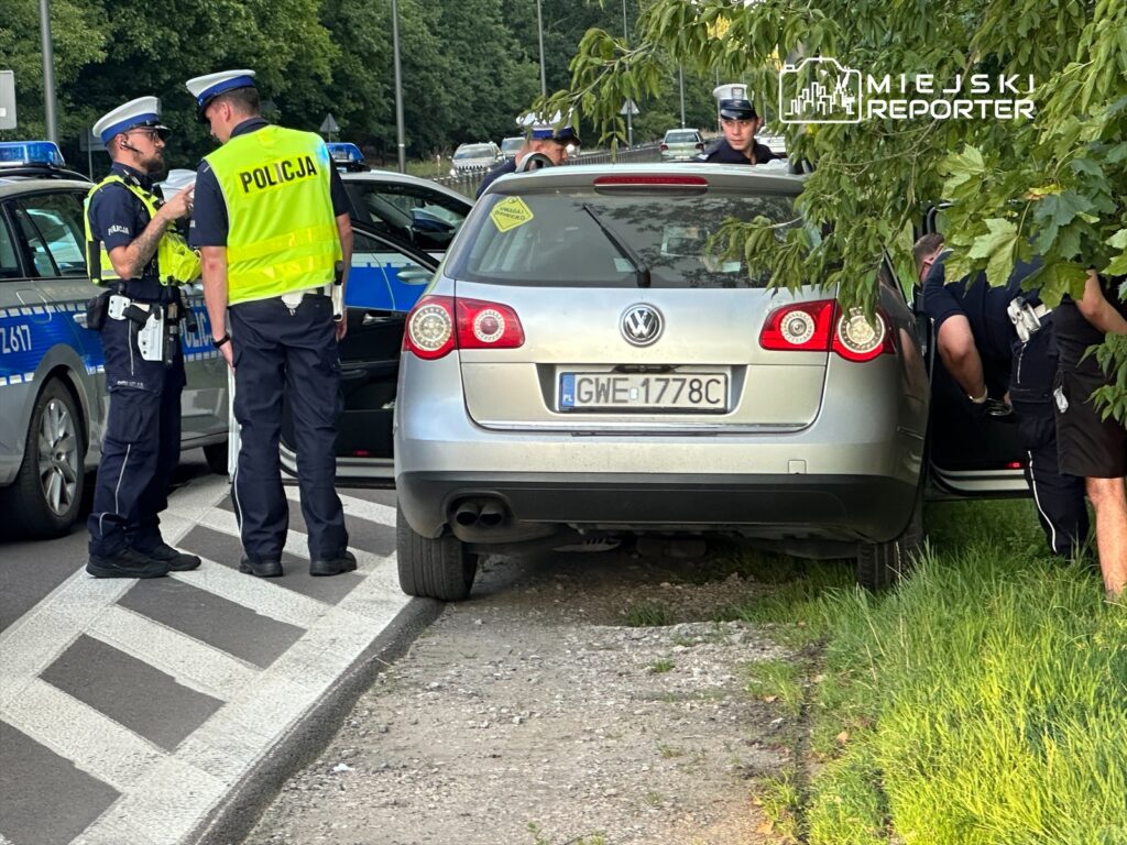 Dwóch policjantów rozmawia obok zatrzymanego samochodu osobowego na poboczu drogi, w tle inne radiowozy.