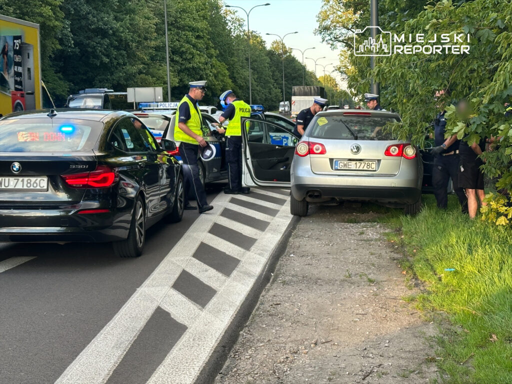 Policjanci kontrolują kierowcę przy zaparkowanym na poboczu samochodzie osobowym w miejskim otoczeniu.