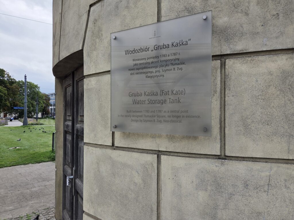 Tablica informacyjna opisująca Wodociąg "Gruba Kaśka" umieszczona na budynku, w tle zieleń i chodnik.