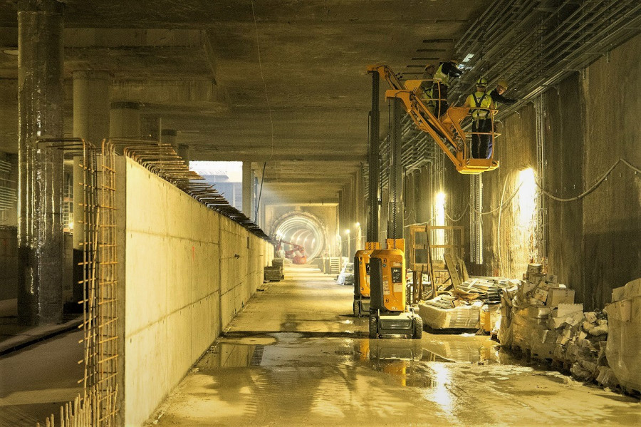 Pracownicy w kamizelkach ochronnych montują instalacje na wysokości w podziemnym tunelu budowlanym.