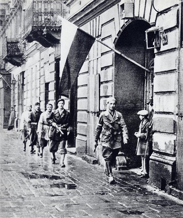 Grupa żołnierzy w mundurach idzie wzdłuż ulicy, obok budynku z flagą wietrzącą na wietrze.