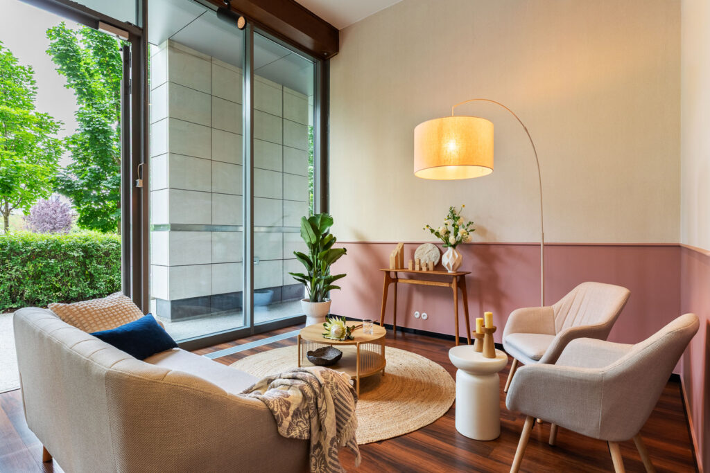 Nowoczesny salon z dużymi oknami, jasną sofą, drewnianą podłogą i okrągłym dywanem, z rośliną i lampą podłogową.