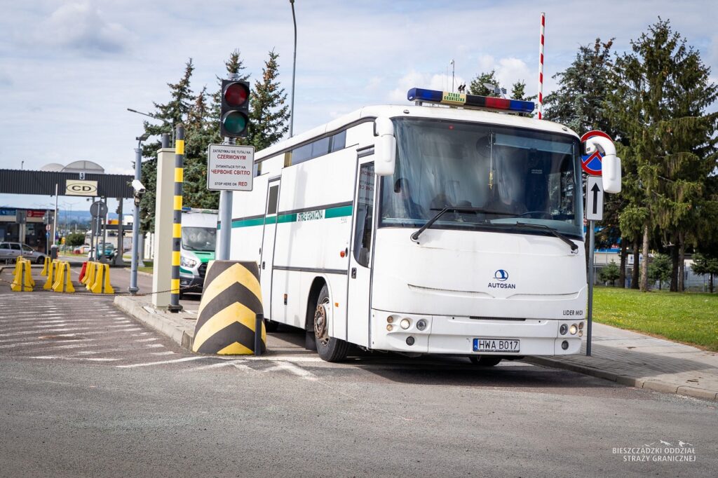 Biały autobus Straży Granicznej zatrzymany na czerwonym świetle przy przejściu granicznym, otoczony barierkami.