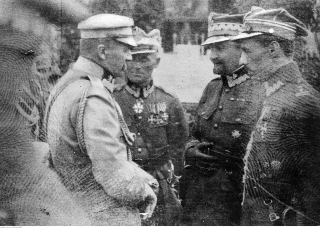 Czterech mężczyzn w mundurach wojskowych rozmawia w plenerze, przy dekoracjach i drzewach w tle.