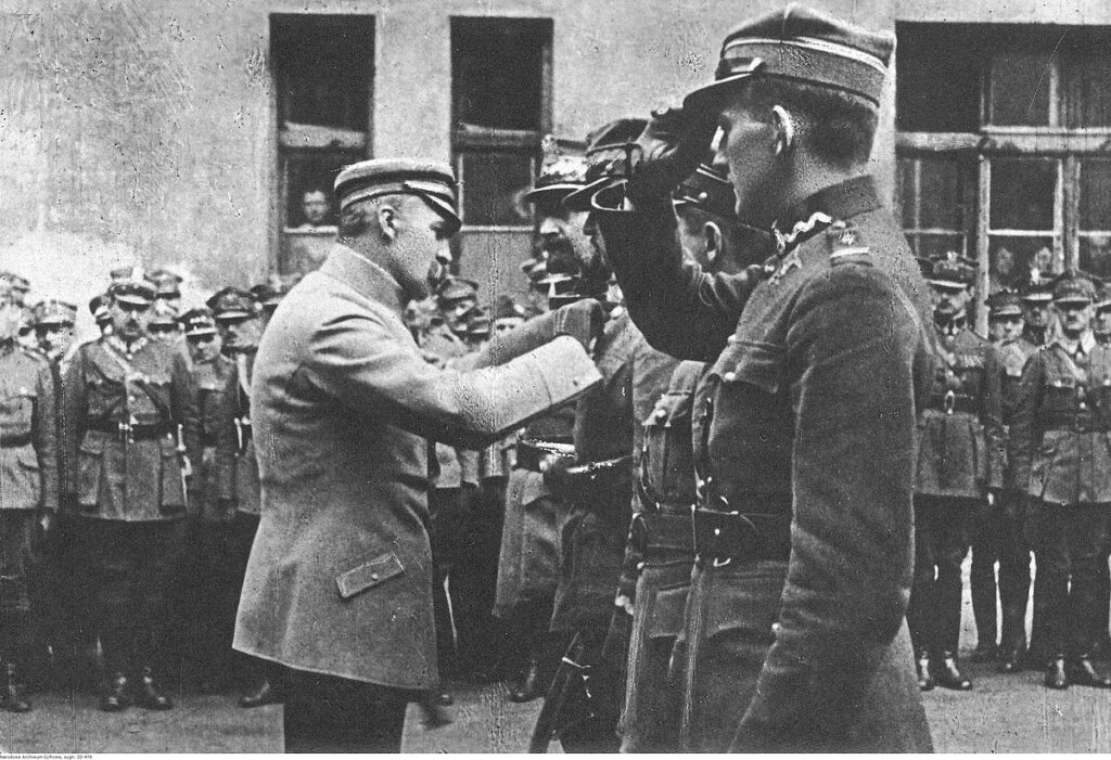 Mężczyzna w mundurze wojskowym odbierający odznaczenie od oficera w garniturze przed grupą żołnierzy.