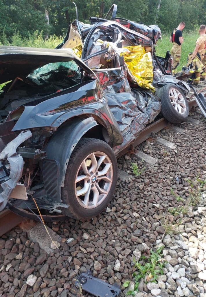 Zniszczony samochód osobowy leży na torach kolejowych, otoczony przez trawę i kamienie, obok stoją strażacy.
