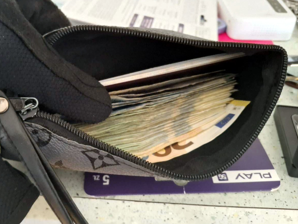 Ręka w czarnej rękawiczce trzyma otwartą torebkę, w której widoczne są banknoty oraz dokumenty.