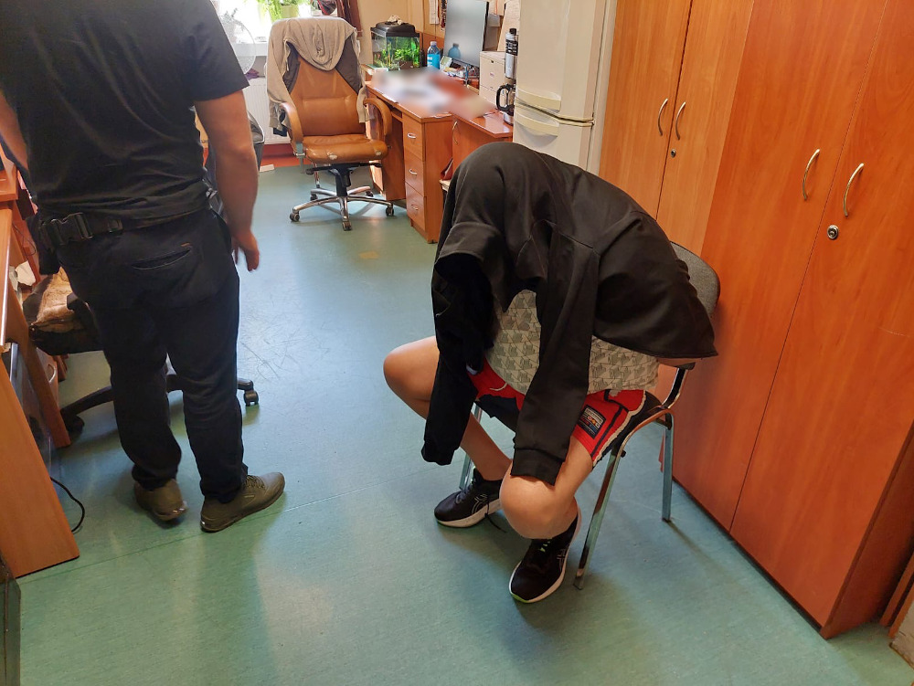 Mężczyzna w czarnej bluzie siedzi na krześle z głową opartą na kolanach, a obok stoi inny mężczyzna w ciemnych ubraniach.