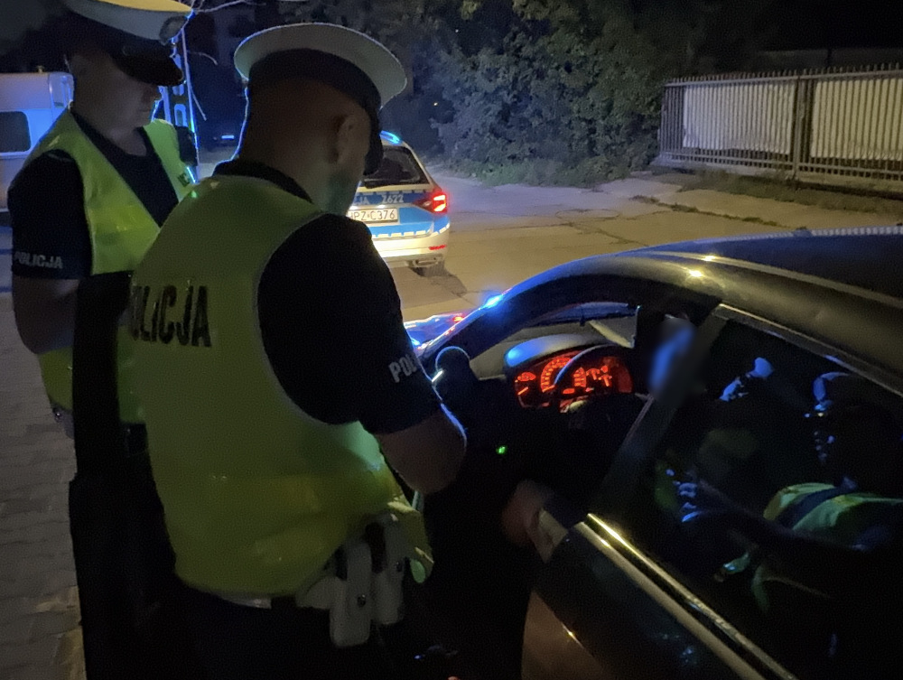 Dwaj policjanci w odblaskowych kamizelkach kontrolują kierowcę w samochodzie nocą. W tle widać radiowóz.