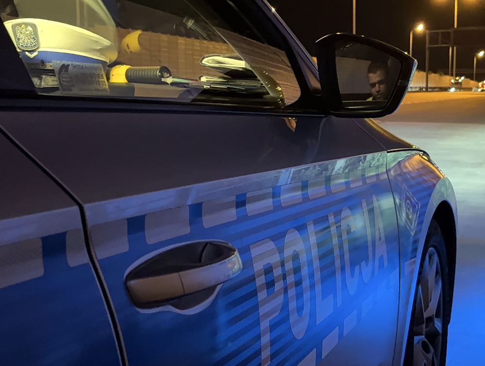 Policyjny samochód zaparkowany na nocnej ulicy, z widocznym napisem "POLICJA" na drzwiach.
