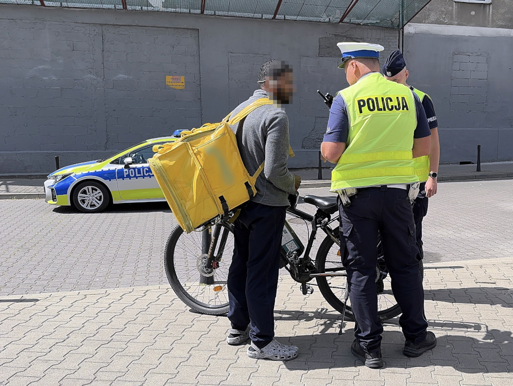 Mężczyzna z żółtą torbą na rowerze rozmawia z policjantami na ulicy, obok radiowozu z napisem "POLICJA".