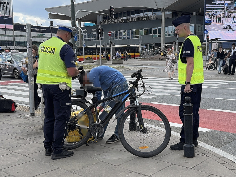 Mężczyzna przy rowerze zostaje kontrolowany przez dwóch policjantów na ruchliwej ulicy w pobliżu dworca.