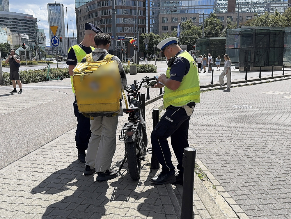 Trzech policjantów rozmawia z mężczyzną na rowerze dostawczym w miejskim otoczeniu, obok nowoczesnych budynków.