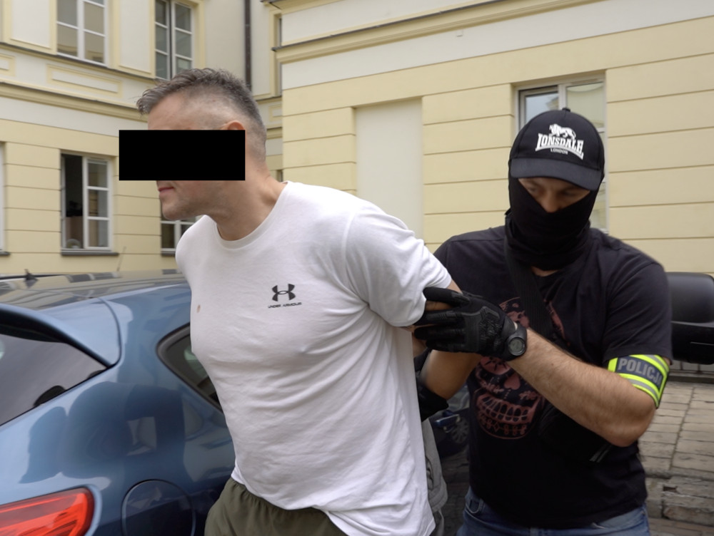 Mężczyzna w białej koszulce jest zatrzymywany przez policjanta w czarnej masce na ulicy, obok samochodu.