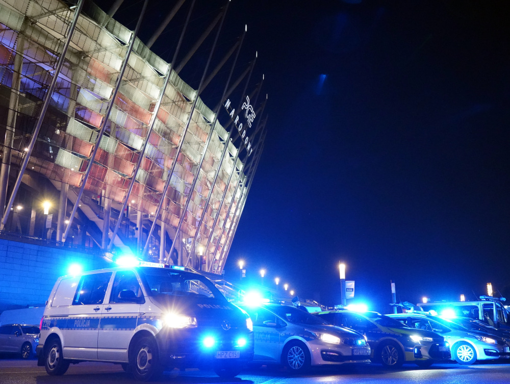 Policyjny radiowóz z włączonymi sygnałami świetlnymi stoi przed nowoczesnym stadionem, oświetlonym w nocy.
