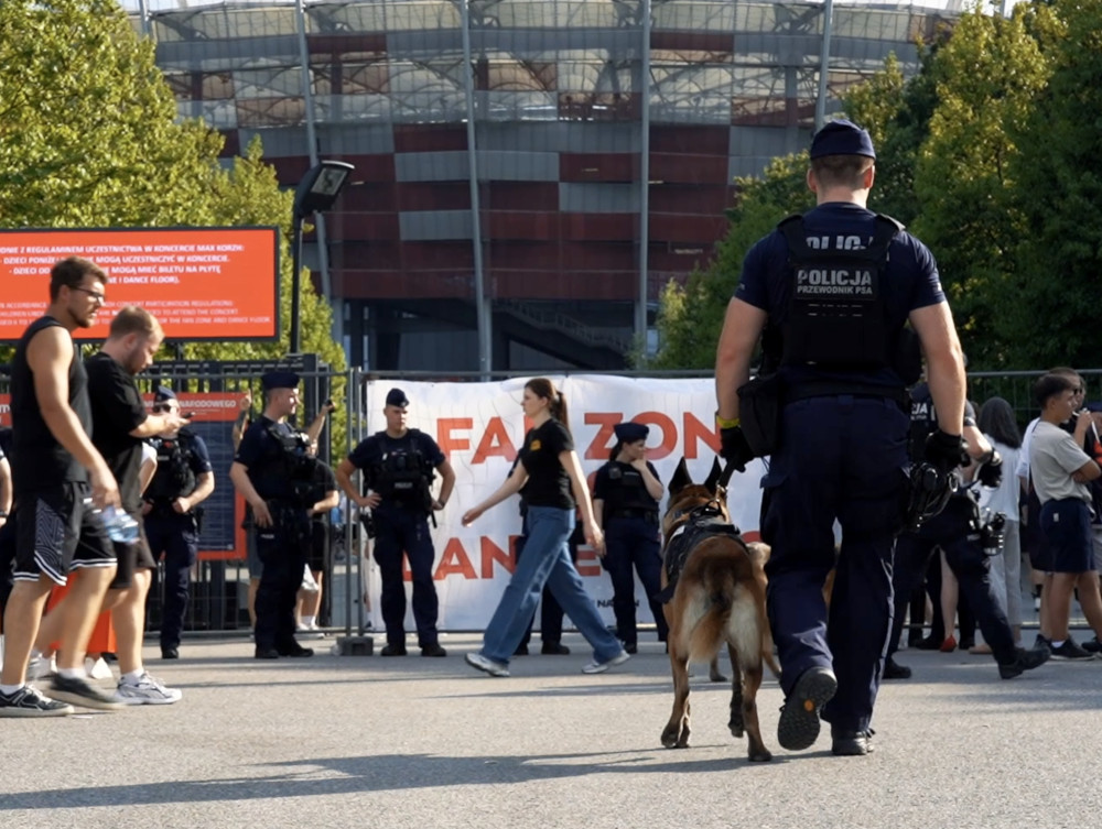 Grupa policjantów z psem służbowym kontroluje tłum przed stadionem, w tle widoczny transparent z napisem.