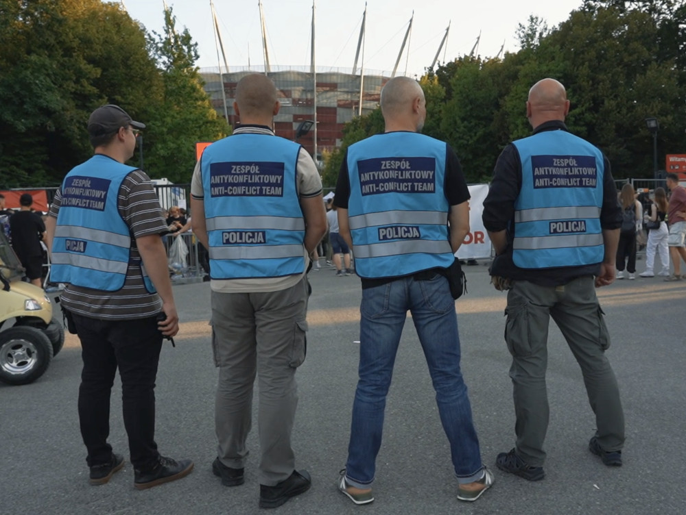 Czterech funkcjonariuszy w kamizelkach z napisem "Zespół Antykonfliktowy" obserwuje tłum przed stadionem.