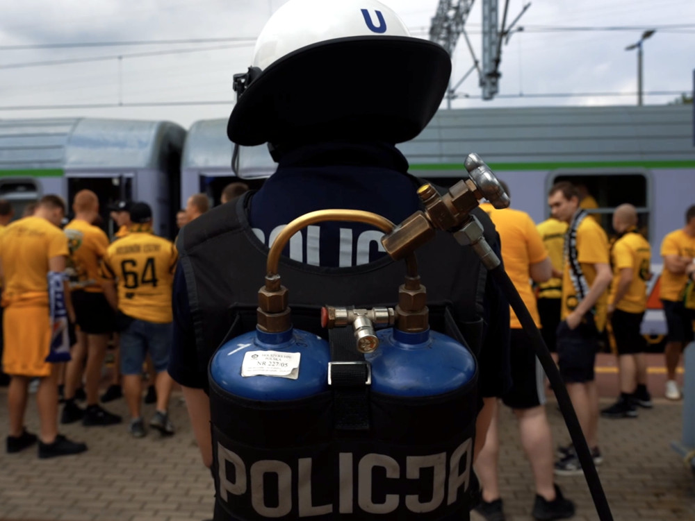 Policjant w hełmie z urządzeniem na plecach obserwuje grupę kibiców w żółtych koszulkach na stacji kolejowej.
