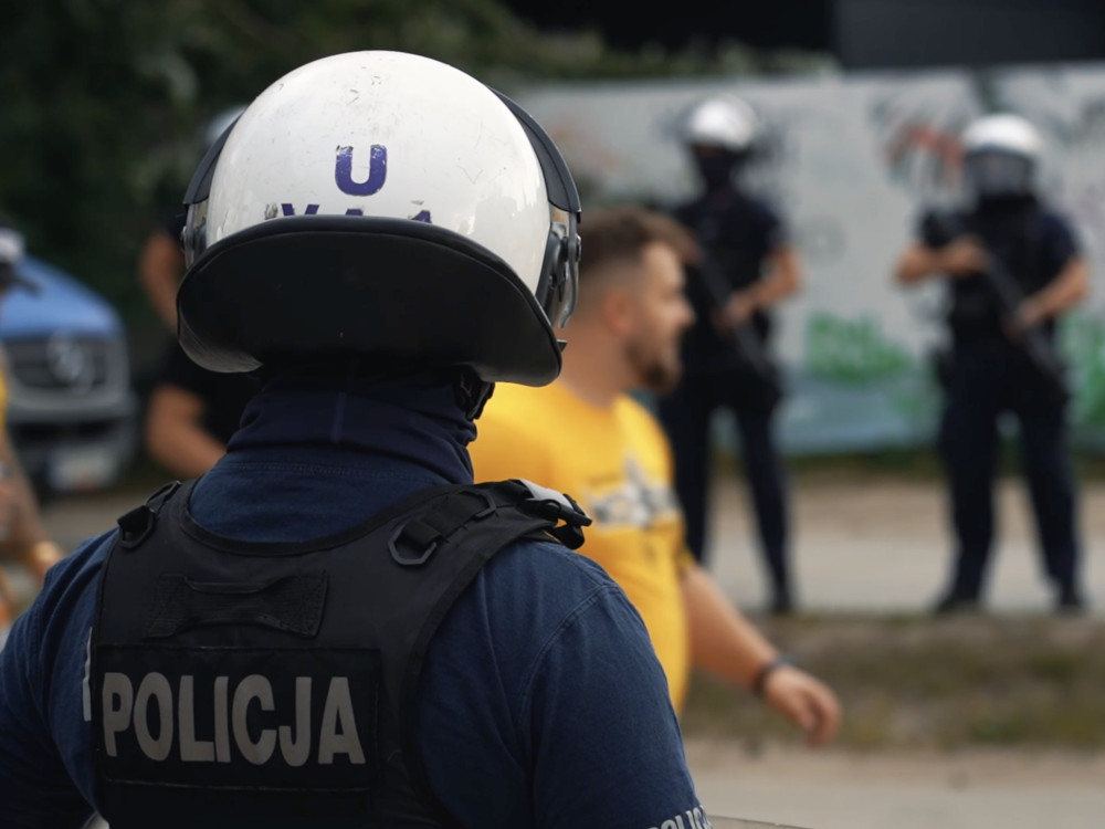 Policjant w hełmie obserwujący grupę ludzi w tle, z kolegami w mundurach trzymającymi broń.