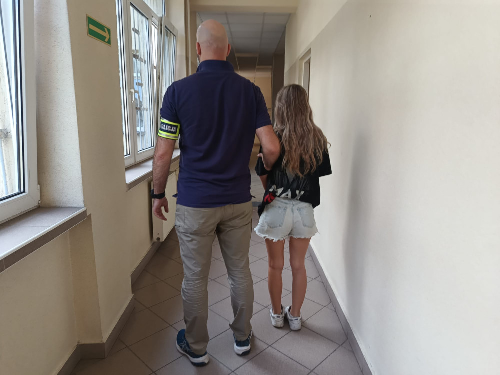 Mężczyzna w mundurze policjanta prowadzi dziewczynę w szortach przez korytarz budynku.