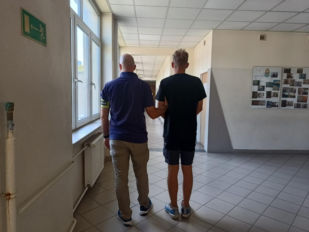 Mężczyzna w mundurze z opaską prowadzi drugiego mężczyznę korytarzem w budynku, obok okien i zdjęć na ścianie.