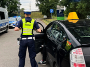Policjant w kamizelce odblaskowej kontrolujący pasażera w czarnym samochodzie na parkingu.