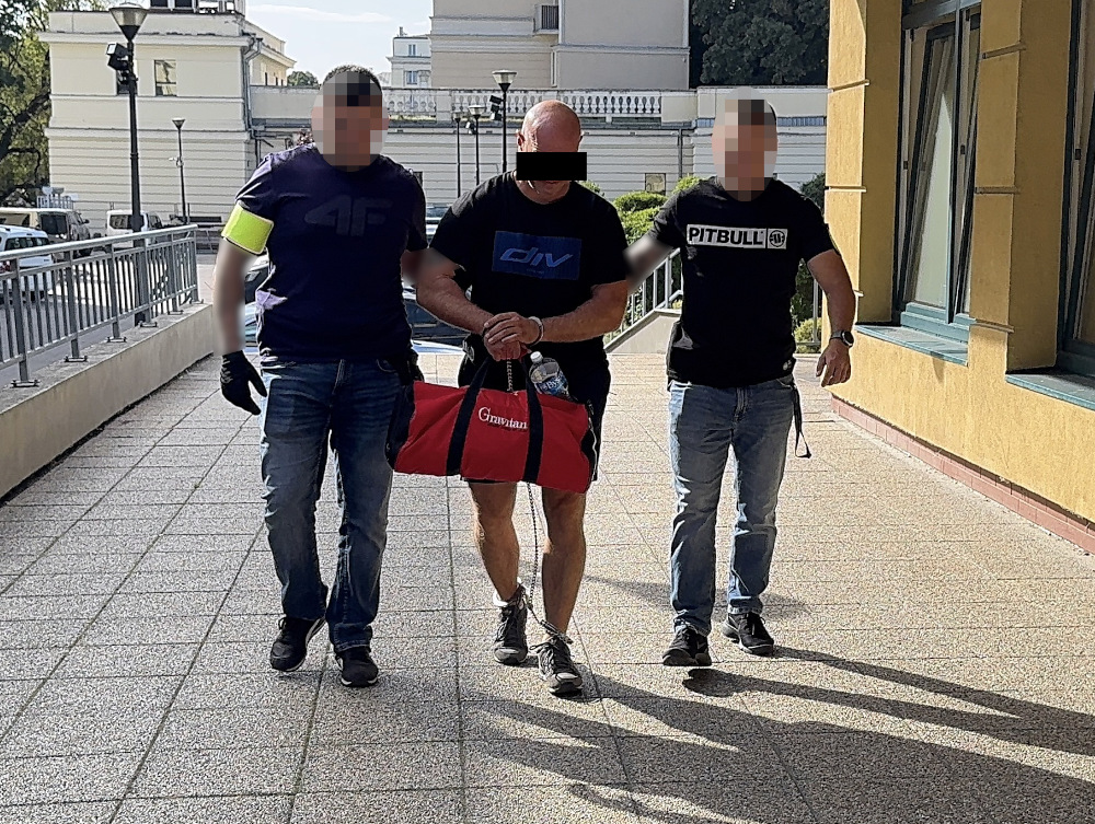 Trzech mężczyzn idących po chodniku, jeden z nich niesie czerwoną torbę, w tle budynki i latarnie uliczne.